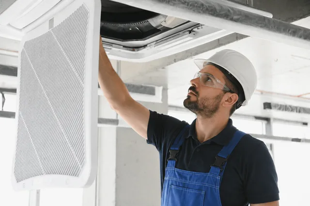 HVAC technician in blue overalls and a white hard hat servicing a ceiling air conditioner up close